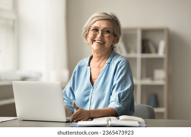 Happy older freelance business lady in stylish glasses working at laptop computer, typing, sitting at home workplace desk, smiling at camera for portrait, enjoying domestic Internet, remote job