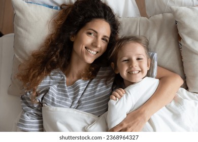 Top view portrait of smiling young mother and cute little daughter lying in bed under blanket ready to sleep, happy millennial mom or nanny hug cuddle in bedroom with small preschooler girl child