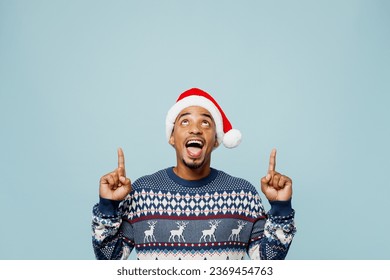 Joven sorprendido con un suéter de punto y un gorro de Papá Noel posando con el dedo índice en la cabeza sobre una maqueta aislada sobre un fondo azul pastel. Concepto de celebración de Navidad y Año Nuevo 2024