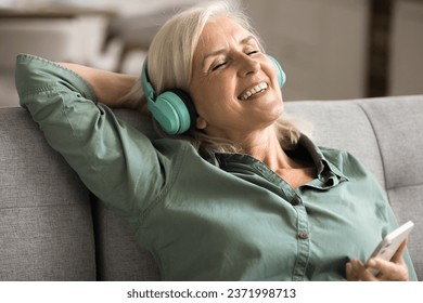 Cheerful inspired blonde elderly woman singing song at home, listening to music from big wireless headphones and online media service on cellphone, resting on sofa with closed eyes and toothy smile