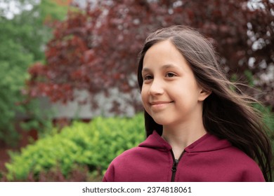 Ritratto di una gioiosa bambina bruna di 10-11 anni con i capelli lunghi, sullo sfondo di foglie rosse e verdi sulla strada.