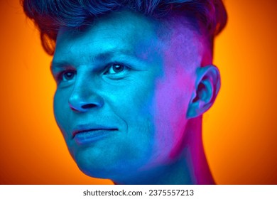 Portret z bliska młodego przystojnego uśmiechniętego mężczyzny, studenta patrzącego w bok na tle gradientowego pomarańczowego tła oświetlonego niebieskim neonowym światłem. Koncepcja piękna, młodości, samorealizacji, ludzkich emocji.