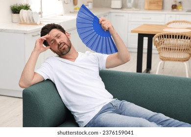 Hombre barbudo agitando un abanico azul para refrescarse en el sofá de su casa