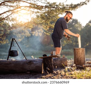 Un homme barbu, un bûcheron avec une hache dans les mains, coupe du bois dans la nature en été avec en toile de fond une forêt et une rive de rivière. Camping. Survie.