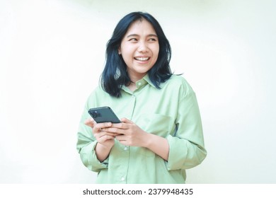Grua e suksesshme aziatike me flokë të errët e veshur me këmishë të gjelbër me madhësi të madhe duke përdorur rrjetet sociale në smartphone duke parë larg e sigurt duke qëndruar mbi sfond të bardhë të izoluar