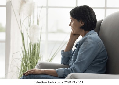 Pensive mature woman sits on armchair staring into distance, deep in thoughts, pondering, thinks on life problems, spend time alone at home, suffering from solitude and boredom on retirement. Concerns