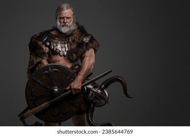 A rugged, silver-haired, bearded elderly viking dressed in fur and lightweight armor, with a helmet attached to his belt, holding an axe and shield against a gray background