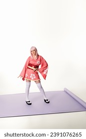Beyaz stüdyoda mor halı üzerinde pembe kimono ve sarı peruklu anime tarzı kadın, eli kalçasında