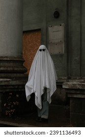 A imagem retrata uma pessoa vestida com uma fantasia branca de fantasma, usando óculos escuros, parada em frente a um pilar.