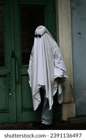 A imagem mostra uma pessoa vestindo uma fantasia de fantasma branca com óculos escuros, parada em frente a uma porta verde.