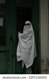 A imagem mostra uma pessoa usando uma fantasia de fantasma branca e óculos de sol, parada em frente a uma porta verde com uma placa que diz "fechado".