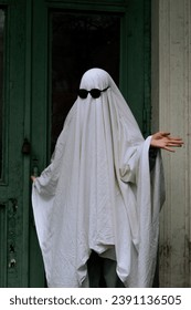 A imagem mostra uma pessoa vestida com uma fantasia de fantasma branca e óculos de sol pretos, parada em frente a uma porta verde.