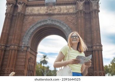 Attraktiv jonk weiblech Tourist entdeckt nei Stad. Fra hält eng Pabeierkaart op zentrale Fra mécht Selfie géint erstaunlech Architektur vum Arc de Triomphe Gate zu Barcelona