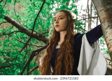 Portret wojowniczki-elfki w niebieskiej sukni i rudych włosach w lesie.