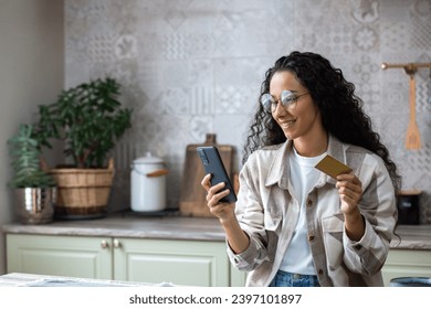 Young beautiful woman in kitchen with phone and bank credit debit card in hands, smiling Hispanic woman chooses gifts and products in online store, online shopping remotely from home.