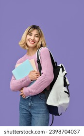 Glückliche, hübsche, blonde Studentin der Generation Z mit kurzen blonden Haaren, die einen Rucksack mit Notizbüchern hält und einen rosa Jeanspullover trägt, der in die Kamera schaut und isoliert auf violettem Hintergrund steht. Vertikales Porträt.
