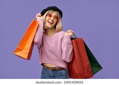 Glückliches, blondes, lächelndes Mädchen der Generation Z, stilvolle junge Frau mit einer Sonnenbrille in der Farbe eines rosa Pullovers und Einkaufstüten, zufrieden mit Modeeinkäufen zu Sonderpreisen, isoliert auf violettem Hintergrund.