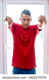 Portrait of a handsome young man showing thumb down indoors, standing next to window. No way