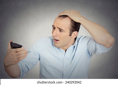 Closeup portrait shocked man feeling head, surprised he is losing hair, receding hairline or seeing bad news on cell phone isolated grey wall background. Negative facial expressions, emotion feeling