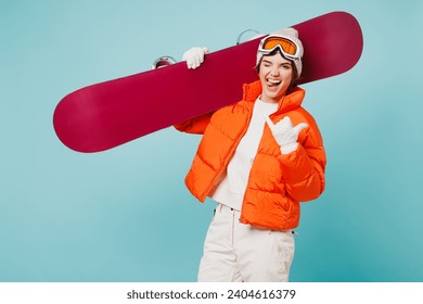 Tânără femeie fericită purtând o jachetă căptușită caldă, cu căptușeală, ochelari de schi, mască, ține snowboard, arată, shaka, gest, clipește, călătorie, odihnă, petrece weekendul, sezonul de iarnă în munți, izolat pe fundal albastru simplu
