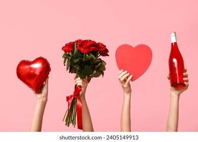 Female hands holding roses, bottle of wine, heart-shaped balloon and gift box on pink background. Valentine's Day celebration