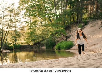 Happy, energetic girl in white t-shirt and black leggings running on sandy beach close to river. Little athlete with dark long hair doing sports and having fun in forest near water.