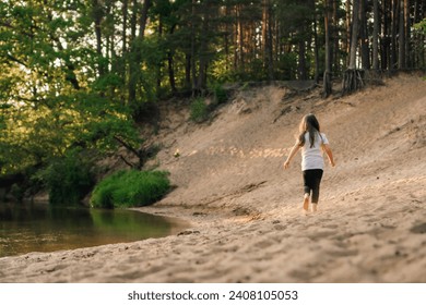 Rear view of little girl with dark, long and flowing hair running on sandy beach near river in forest. Female child in white t-shirt and black leggings actively spending time in nature.