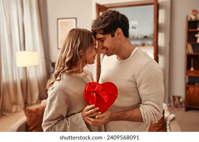 Happy Valentine's Day. A man gives a heart-shaped gift box to his beloved woman in the living room at home, the woman hugging him tenderly. Romantic evening together.