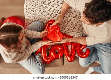 Happy Valentine's Day. Young couple in love exchanging gifts while sitting on the sofa in the living room at home. A man giving a heart-shaped gift box to his beloved woman.