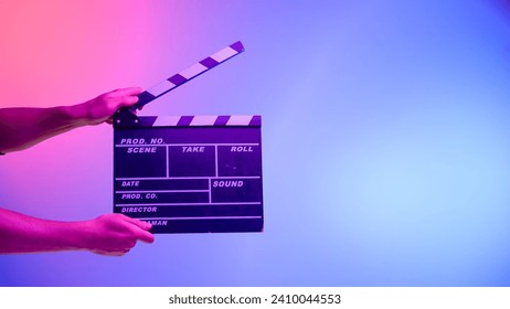 Hand hält schwarze Filmklappe oder Filmtafel auf blauem Hintergrund mit kreisförmigem Licht, beleuchtet mit rosa Neonlicht. Kinoindustrie, Unterhaltung.