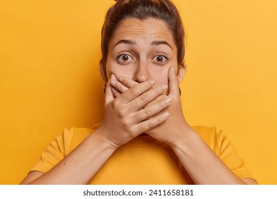 Tânăra femeie drăguță își acoperă gura cu ambele mâini încearcă să nu lase niciun cuvânt se uită impresionat încearcă să rămână fără cuvinte îmbrăcată în tricou casual izolat pe fundal galben gâfâie de uimire