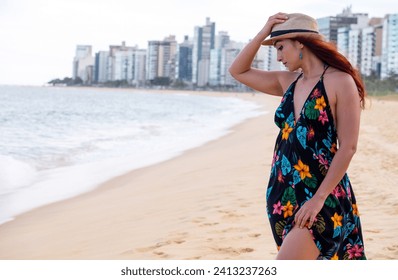 Žena u haljini i šeširu bezbrižno hoda po pijesku plaže sa gradom u pozadini. Osvetljenost i kontrast odevne kombinacije sa pejzažom prenose opuštenost i spokoj.