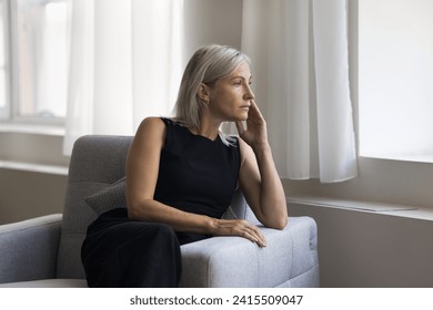 魅力的な成熟した 50 代のブロンドの女性が、居心地の良いアームチェアに座って一人でくつろぎ、窓の外を見つめながら考え事をし、夫を待ち、行方不明になり、深く考え込み、問題の解決策を探し、物思いにふけり、思慮深く見える。