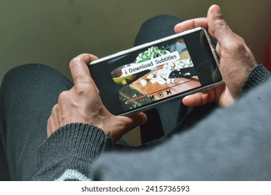 Man watching a cooking show on his smartphone with the message download Subtitles. Get subtitles for different language translation concepts. Selective focus.