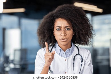 Médico afroamericano serio con bata blanca levantando el dedo índice con expresión facial rigurosa. Cardiólogo experimentado que enfatiza los daños que causa el tabaquismo al sistema cardiovascular de los adultos.