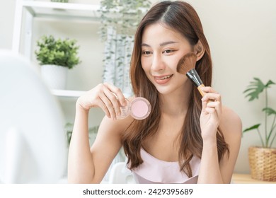 Happy beauty blogger concept, cute asian young woman, girl smile, make up face by applying brush blush powder on her cheek, looking at the mirror. People look with natural fashion style.