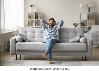 Carefree wellbeing barefoot Indian guy enjoy fresh conditioned air leaned on comfortable sofa in fashionable living room, relaxing alone with hands behind head. Rest, leisure, daydreams, smart home