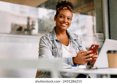 Una joven alegre disfruta de un descanso para tomar café en una cafetería. Utiliza su teléfono móvil para comunicarse y navegar por Internet. Con una sonrisa sincera, interactúa con las redes sociales y el aprendizaje electrónico.