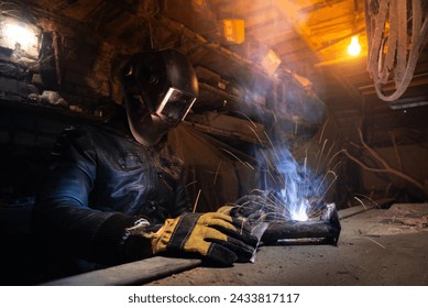 Young mechanic with a welding machine in an old dirty garage at night. Man in protection doing welding work, hobby