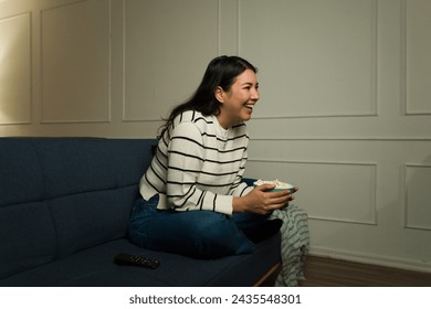 امرأة آسيوية متحمسة تضحك وتستمتع أثناء مشاهدة برنامج تلفزيوني أو فيلم كوميدي أثناء تناول الفشار على الأريكة