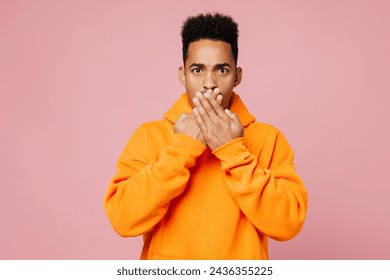 Jonk traureg verréckten Mann vun afroamerikanesch Ethnie hien huet giel Hoodie Casual Kleeder Cover Mond mat Hänn kucken Kamera isoléiert op Einfache Pastell hellrosa Hannergrond Studio Portrait. Liewensstil Konzept