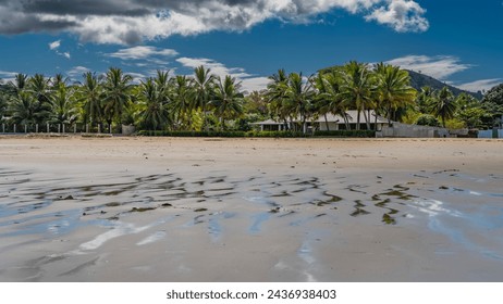 The seabed was exposed at low tide. Streams, puddles of water on the sand. Cottages are visible on the shore, among palm trees.  Clouds in the blue sky. Madagascar. Nosy Be