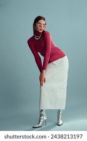 Fashionable confident woman wearing pink sunglasses, turtleneck, layered pearl necklace, white denim maxi skirt, silver boots, posing on blue background. Full-length studio fashion portrait