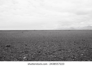 A flat expanse of land covered with volcanic gravel with black sand leading to cloudy mountain in Southern Iceland.