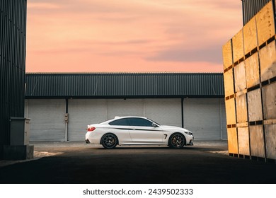 Un'auto sportiva bianca si erge con grazia di fronte a un magazzino usurato mentre il sole tramonta. Immerso nel caldo chiarore del crepuscolo, l'elegante veicolo trasuda eleganza e potenza sullo sfondo industriale