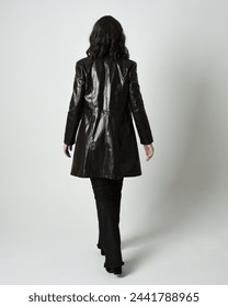 Full length portrait of brunette woman wearing long leather trench coat and black boots. Standing pose walking away from camera, facing backwards. Isolated silhouette on white studio background.