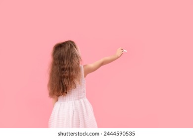 Kleines Mädchen zeichnet mit Kreidestück auf rosa Hintergrund, Rückansicht. Feier zum Kindertag