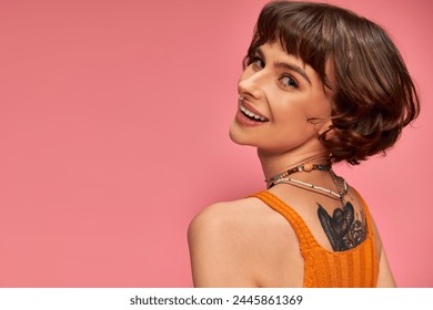 Gelukkige getatoeëerde vrouw in de twintig met neuspiercing en kort bruin haar lachend op roze achtergrond