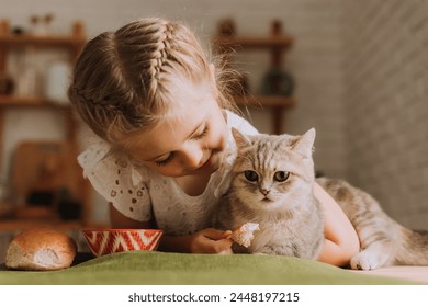 Seorang gadis kecil yang lucu di dapur memberi makan seekor kucing di meja makan. Foto berkualitas tinggi