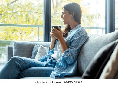 Relaxează-te și relaxează-te. Tânără femeie brunetă frumoasă, cu expresie facială fericită, singură pe canapea, petrecându-și bine cafeaua sau ceaiul în ceașcă. Portret de femeie relaxată care se odihnește acasă. Timp liber